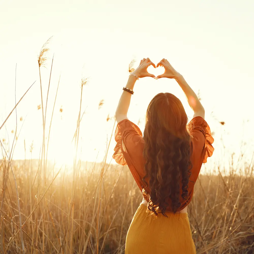 Coaching de vie sur la confiance en soi : une femme de dos forme un cœur avec ses mains face au soleil, symbole d’estime de soi, de force intérieure et de sérénité.