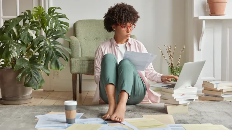Femme assise au sol entourée de papiers et de livres, travaillant sur un ordinateur portable. Illustration du profil Ennéagramme Type 5, l’Observateur, reconnu pour son besoin de compréhension et de réflexion.