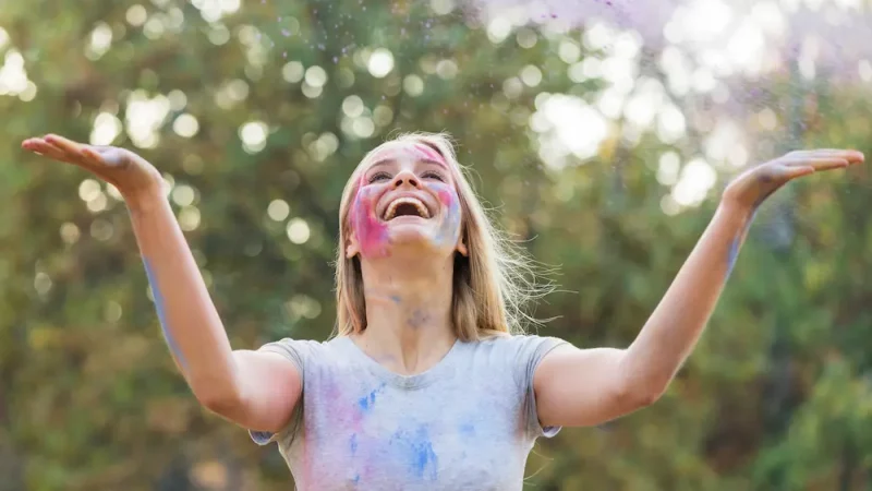 Femme souriante couverte de poudre colorée levant les bras vers le ciel, symbole de joie de vivre et d’énergie positive, illustration du type 7 l’épicurien ennéagramme.
